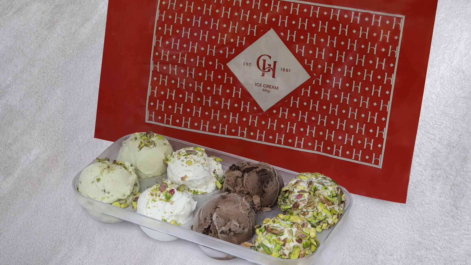 AL HALLAB ice cream sharing box displaying scoops of ashta, chocolate and mixed flavours topped with pistachios, placed in front of branded AL HALLAB ice cream packaging.