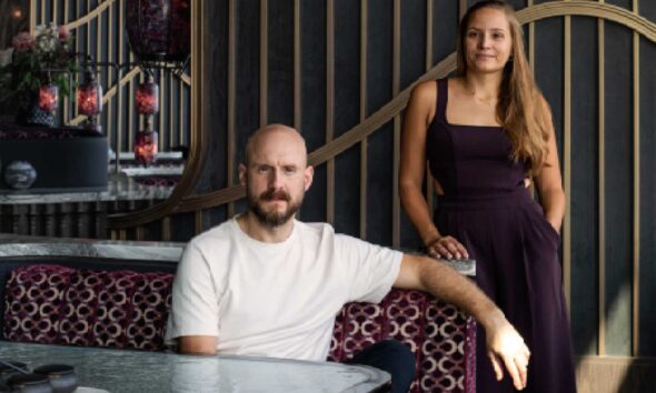 Interior of a Studio WYZE–designed hospitality space featuring two people posed beside a curved banquette, with sculptural vertical wall panels, patterned upholstery, a marble table in the foreground, and layered floral arrangements in a moody, refined setting.