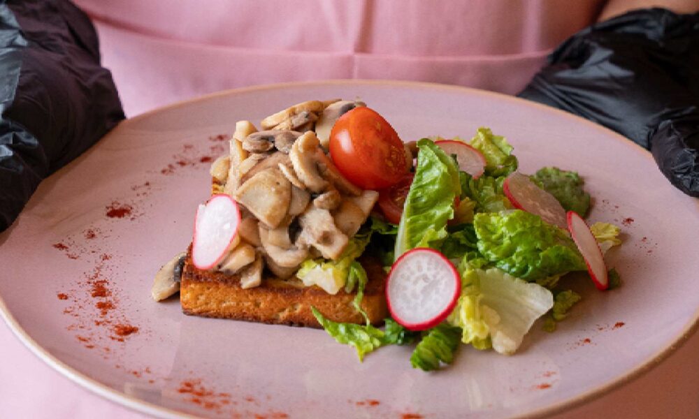 Split image showing Hola Zero Sugar Cafe branding: on the left, a server wearing a Hola Zero apron holding a plated keto dish with vegetables; on the right, the pastel pink Hola Zero Sugar Cafe seating area with diners under decorative floral lighting. ``