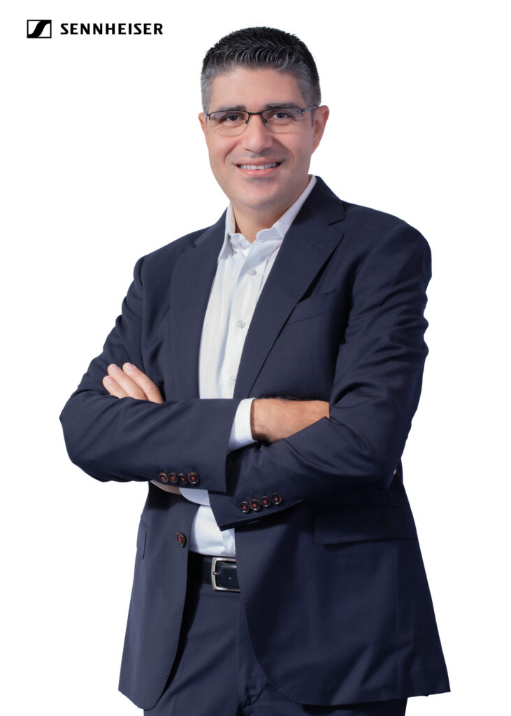 A professional studio headshot of Fadi Costantine, Sales Manager for Business Communication (Middle East) at Sennheiser. He is smiling and standing with his arms crossed against a plain white background. He has short, salt-and-pepper hair, wears glasses, and is dressed in a dark navy blue suit with a white collared shirt. The Sennheiser logo is visible in the top left corner.