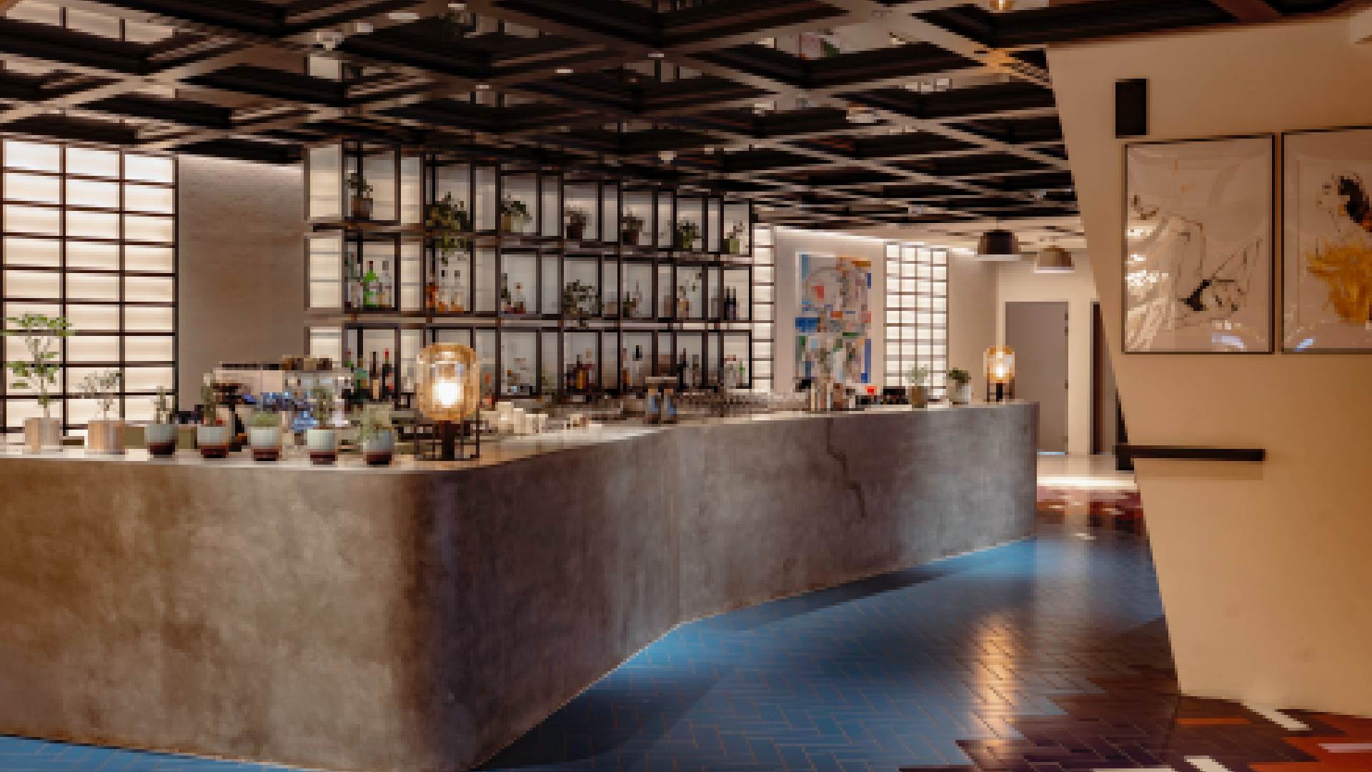 Modern indoor bar at Meliá Dubai featuring a sleek concrete counter, backlit shelving with bottles and plants, warm ambient lighting, and contemporary artwork on the walls