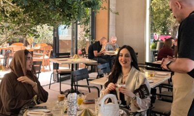 Outdoor dining area at Ergon Agora Dubai featuring a table set with drinks, cutlery, and menus, surrounded by modern black chairs and greenery, with a staff member standing near the table and other guests seated in the background.