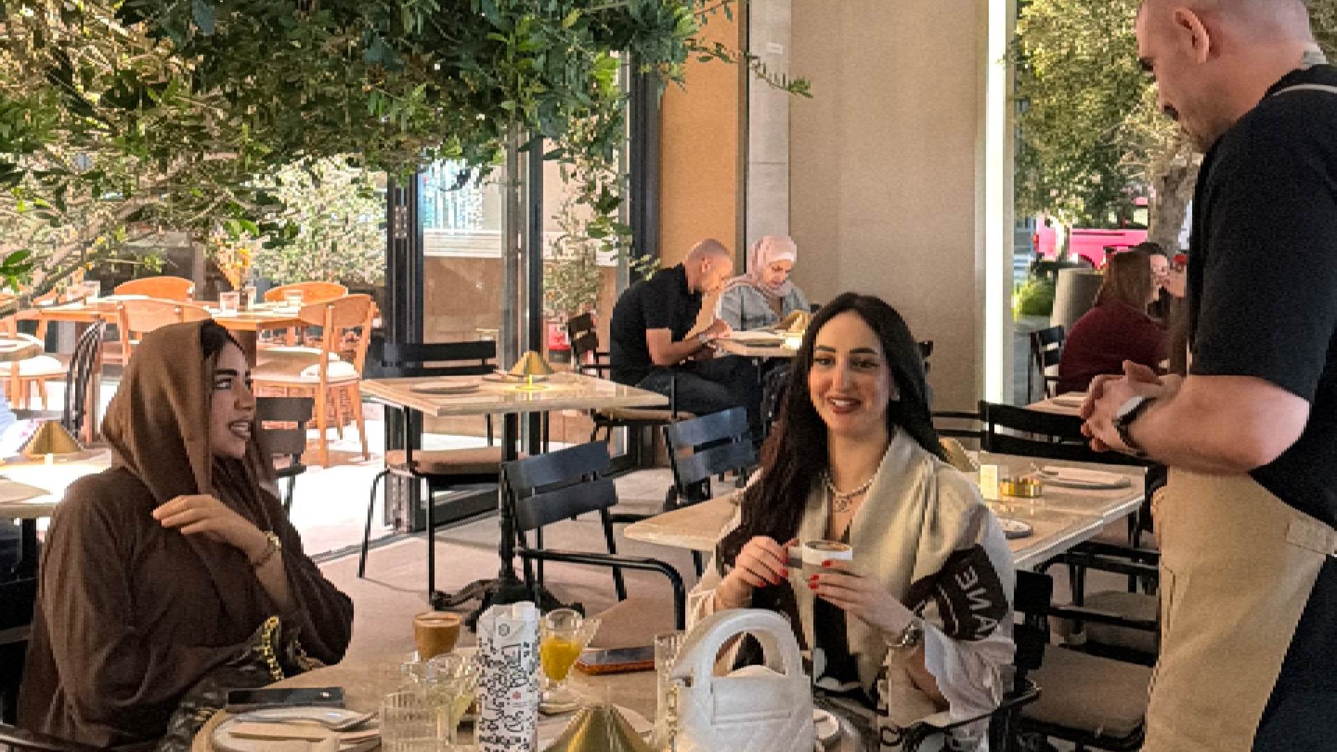 Outdoor dining area at Ergon Agora Dubai featuring a table set with drinks, cutlery, and menus, surrounded by modern black chairs and greenery, with a staff member standing near the table and other guests seated in the background.