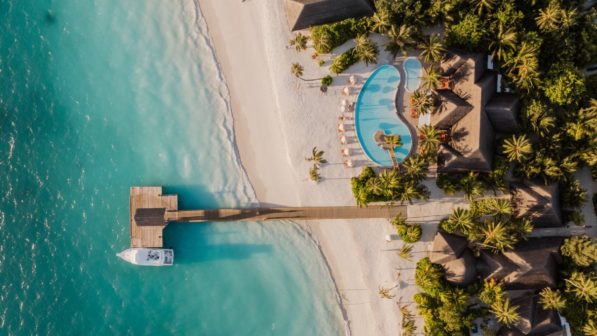 Aerial view of Sun Siyam Iru Veli resort showing turquoise ocean, white sandy beach, wooden pier with a docked boat, and a lagoon-shaped swimming pool surrounded by sun loungers and tropical palm trees. Would you like me to also provide an SEO-friendly caption and optimized image file name for WordPress upload?