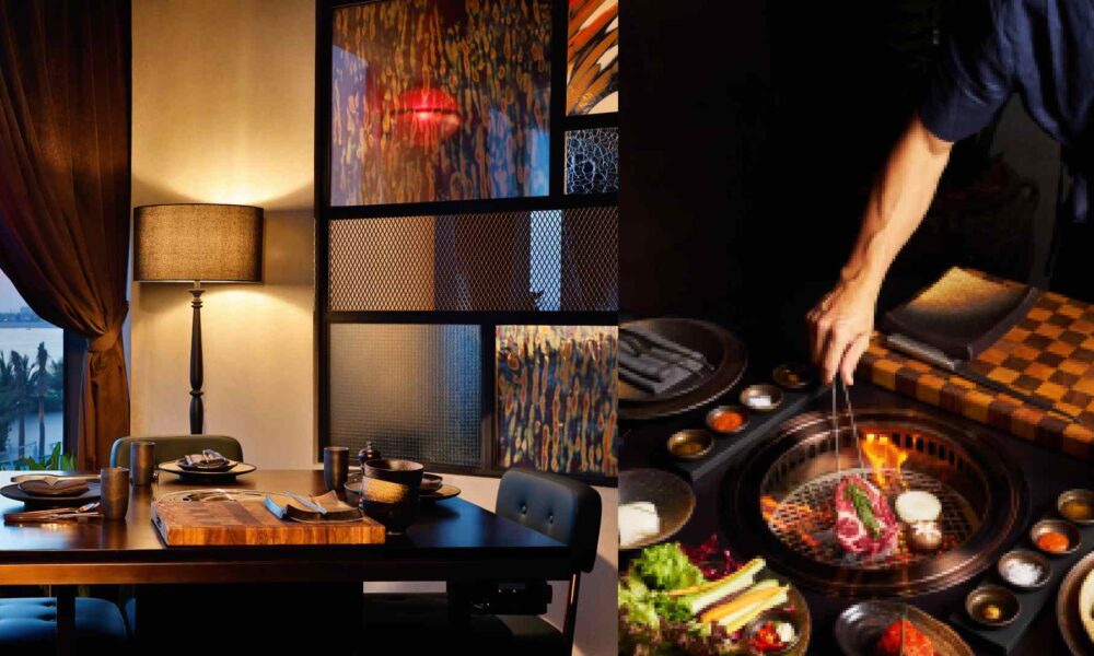 A dining setup at Smoki Moto with a wooden table set for two beside a warm lamp and patterned glass panels, alongside a chef grilling Wagyu meat over an open flame on a Korean barbecue grill surrounded by assorted side dishes and vegetables