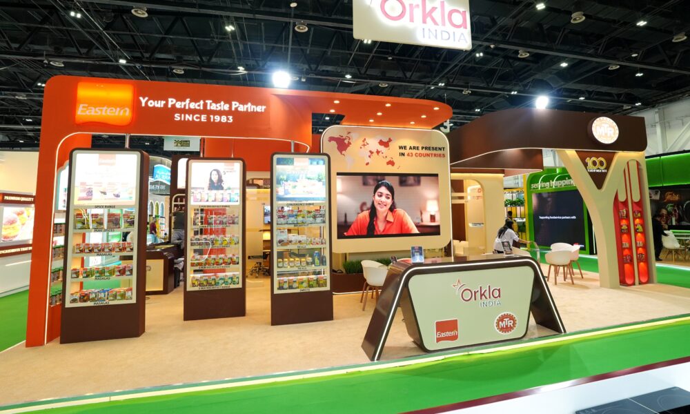 A wide shot of the Orkla India exhibition booth at Gulfood 2026. The vibrant booth features orange and brown accents, showcasing brands like Eastern and MTR. It includes digital displays, product shelves, a world map highlighting presence in 43 countries, and a central reception desk under the Orkla India signage.