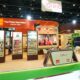 A wide shot of the Orkla India exhibition booth at Gulfood 2026. The vibrant booth features orange and brown accents, showcasing brands like Eastern and MTR. It includes digital displays, product shelves, a world map highlighting presence in 43 countries, and a central reception desk under the Orkla India signage.