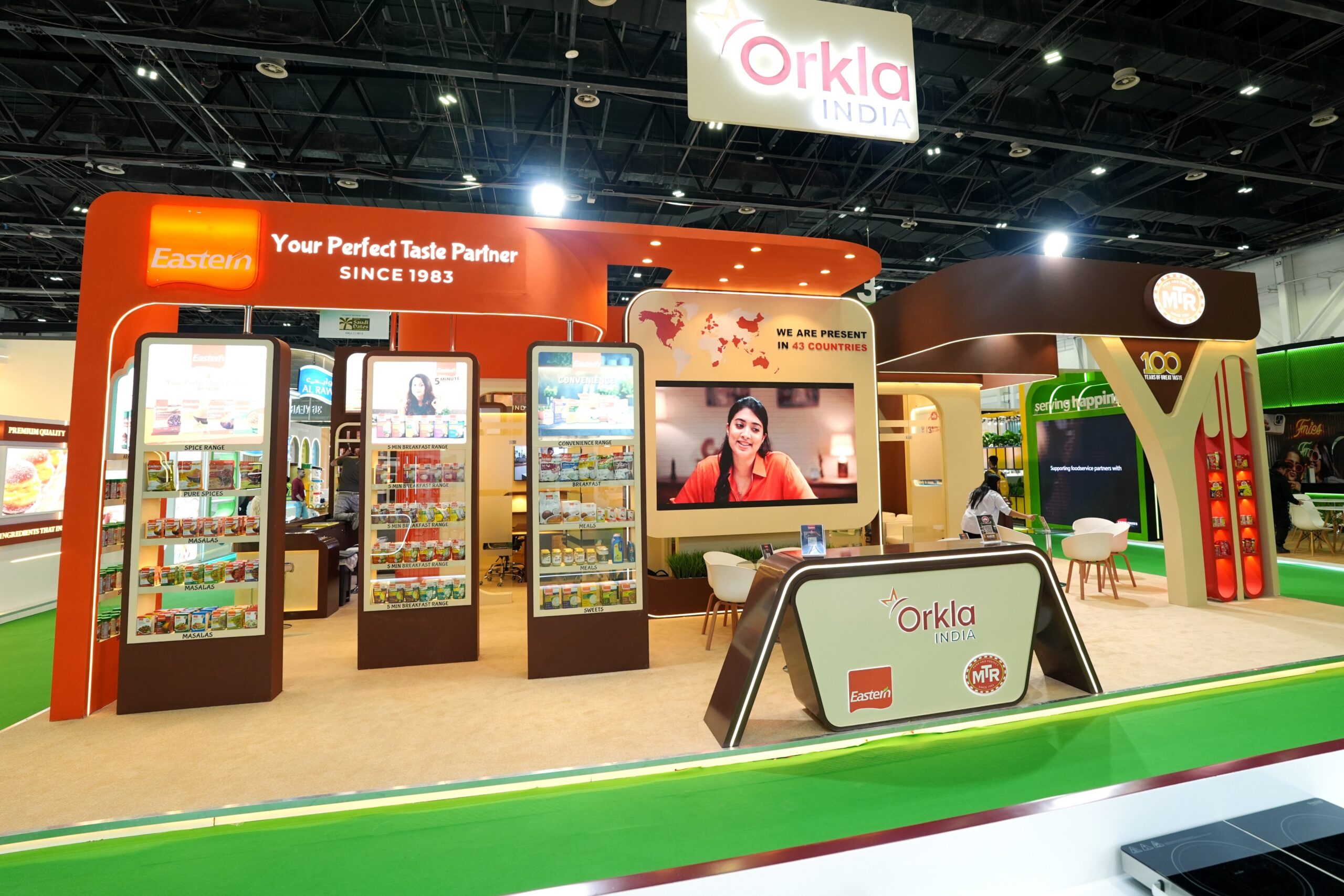 A wide shot of the Orkla India exhibition booth at Gulfood 2026. The vibrant booth features orange and brown accents, showcasing brands like Eastern and MTR. It includes digital displays, product shelves, a world map highlighting presence in 43 countries, and a central reception desk under the Orkla India signage.