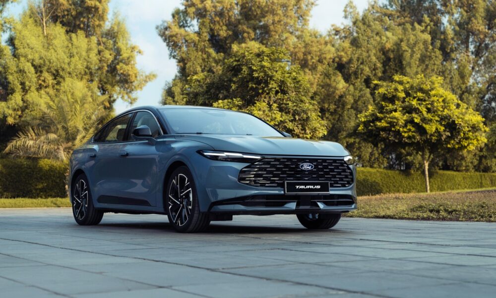 A three-quarter front view of a slate blue Ford Taurus sedan parked on a grey tiled courtyard.