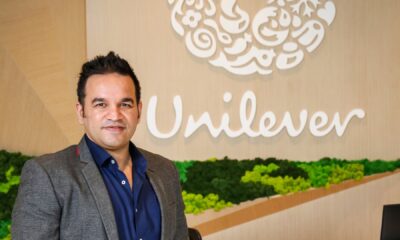 Aseem Puri, CEO of Unilever International, smiling in a professional headshot. He is wearing a grey blazer over a navy blue button-down shirt. Behind him is a light wood-paneled wall featuring the white Unilever logo and a decorative bed of vibrant green moss.