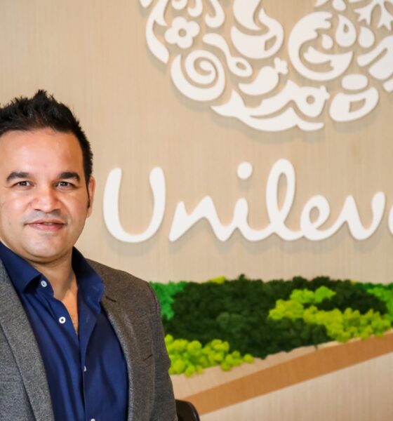 Aseem Puri, CEO of Unilever International, smiling in a professional headshot. He is wearing a grey blazer over a navy blue button-down shirt. Behind him is a light wood-paneled wall featuring the white Unilever logo and a decorative bed of vibrant green moss.