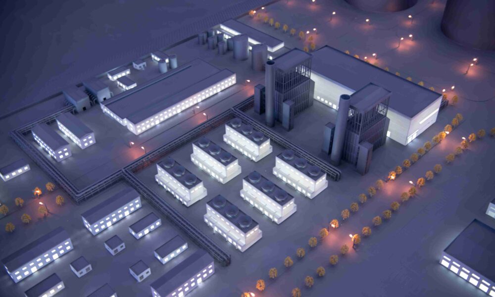 A high-angle, close-up shot of a detailed architectural scale model depicting a large industrial or data center campus at night. The model features various grey buildings with glowing white windows, including long rectangular warehouses and structures with visible rooftop cooling units. Warm orange miniature streetlights line the perimeter roads and small rows of trees, while taller industrial towers with smokestacks stand in the center of the complex.