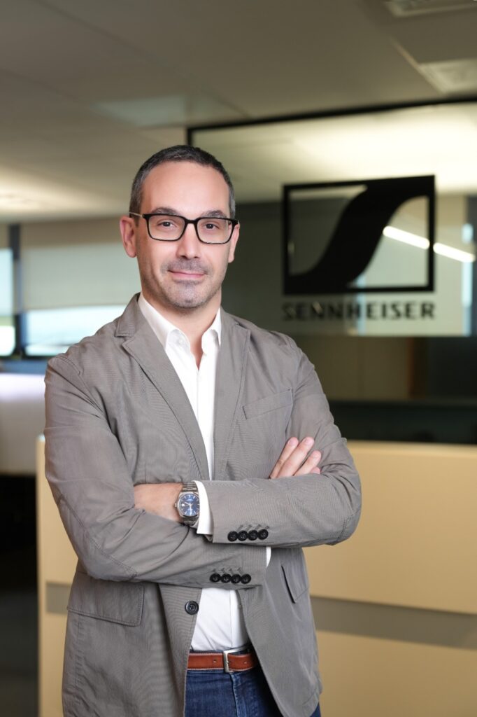 A professional headshot of Mig Cardamone, Sales Director at Sennheiser. He is a middle-aged man with short dark hair and glasses, wearing a grey blazer over a white shirt and jeans. He is standing with his arms crossed in a modern office setting, with a blurred Sennheiser logo visible on the glass wall behind him.