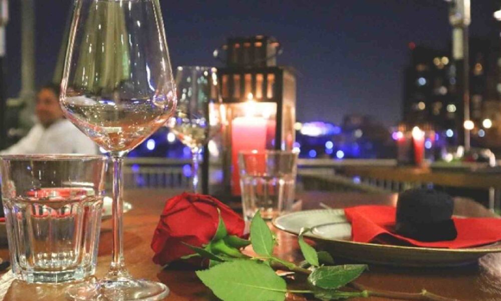 Romantic outdoor dining table set at night with a red rose, wine glasses, water glasses, plates with red napkins, and candles overlooking city lights in the background.