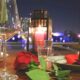 Romantic outdoor dining table set at night with a red rose, wine glasses, water glasses, plates with red napkins, and candles overlooking city lights in the background.