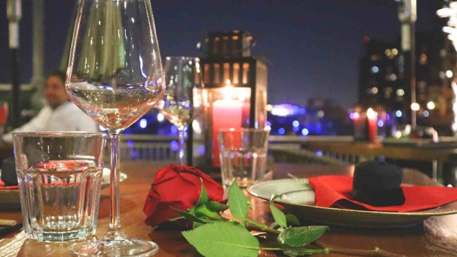 Romantic outdoor dining table set at night with a red rose, wine glasses, water glasses, plates with red napkins, and candles overlooking city lights in the background.