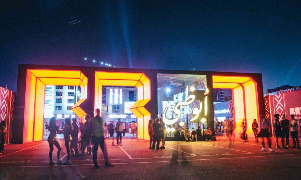 Entrance view of BRED festival at night showing illuminated orange BRED letter structures, digital Arabic calligraphy screen, and attendees walking and gathering at the venue.