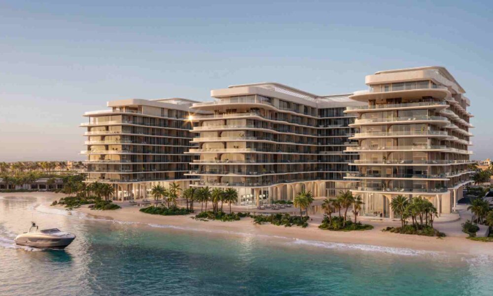 A modern beachfront residential development by Deyaar Development featuring multiple mid‑rise glass‑fronted buildings overlooking a calm shoreline, with landscaped palm trees along the sandy beach and a boat approaching on the turquoise water at sunset.