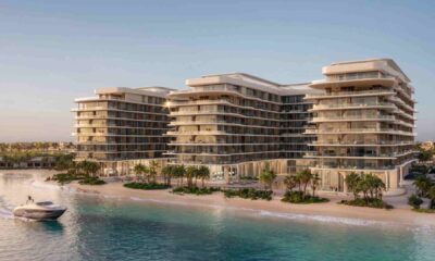 A modern beachfront residential development by Deyaar Development featuring multiple mid‑rise glass‑fronted buildings overlooking a calm shoreline, with landscaped palm trees along the sandy beach and a boat approaching on the turquoise water at sunset.