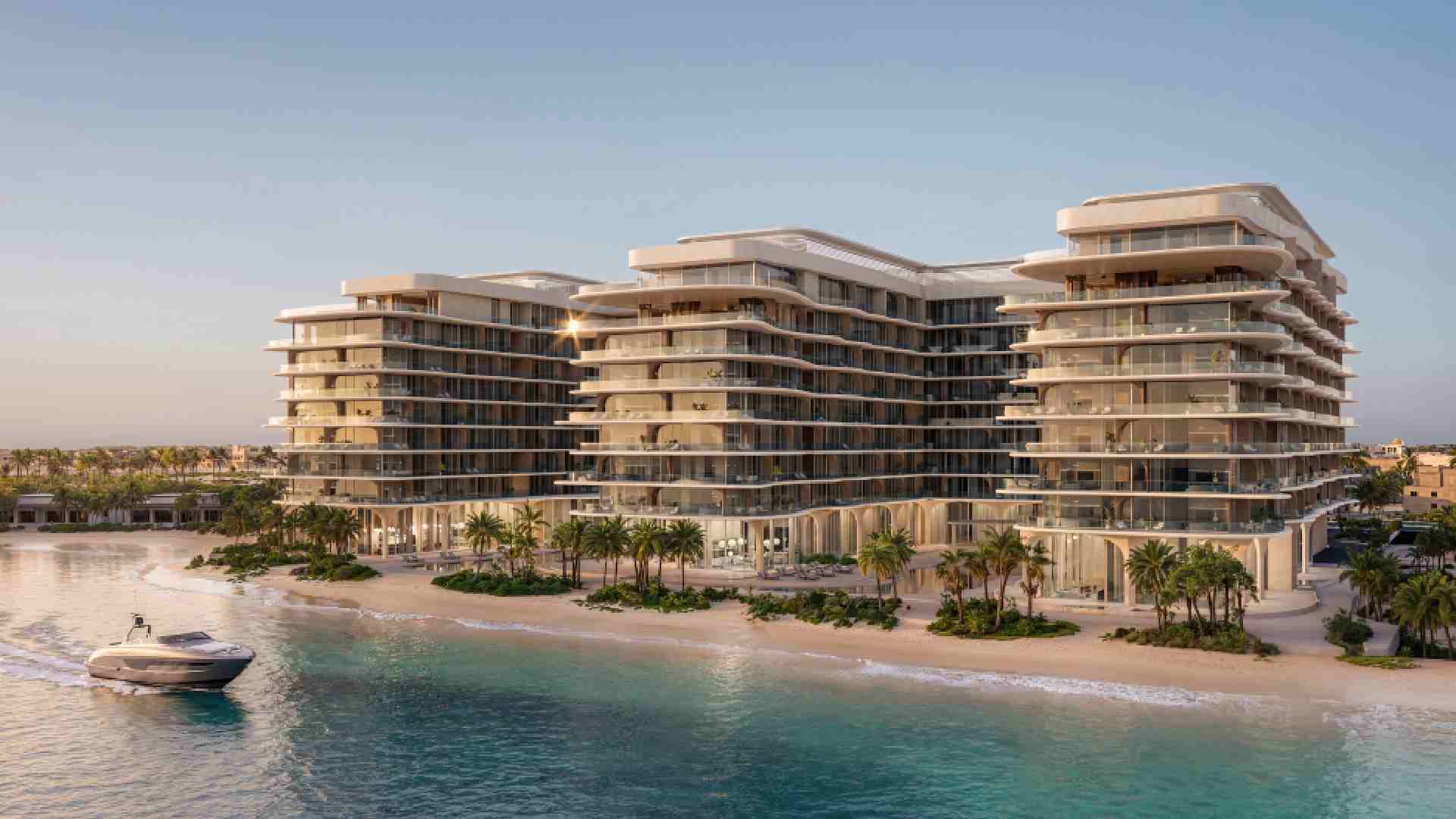 A modern beachfront residential development by Deyaar Development featuring multiple mid‑rise glass‑fronted buildings overlooking a calm shoreline, with landscaped palm trees along the sandy beach and a boat approaching on the turquoise water at sunset.
