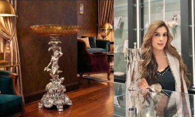 A sculpted silver decorative pedestal with an ornate base and a detailed figurine supporting a large, gold-toned bowl, displayed in a luxurious living room with velvet chairs, wooden flooring, and warm ambient lighting.