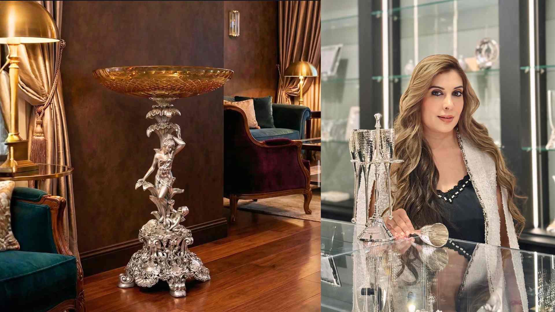 A sculpted silver decorative pedestal with an ornate base and a detailed figurine supporting a large, gold-toned bowl, displayed in a luxurious living room with velvet chairs, wooden flooring, and warm ambient lighting.