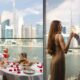 Couple enjoying a private balcony dining experience at InterContinental Residences, seated at a round table with rose petals, cocktails, and plated dishes, overlooking the Dubai Canal and Burj Khalifa skyline, with another scene showing the couple standing by floor-to-ceiling windows clinking glasses against the city view.