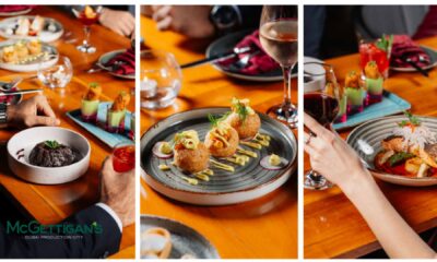Assorted plated dishes and cocktails on a wooden table at McGettigan’s Dubai Production City, with guests seated and dining, showing appetizers, a main course, drinks, and hands reaching for food.