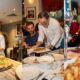 Chefs presenting freshly prepared Italian dishes at LUNAROSSA, featuring a long truffle pizza on a marble counter, a plated pasta dish served at a dining table, and a close-up of a seafood dish with prawns and garnish.