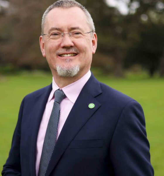 Man in a navy suit and pink shirt wearing an Irish Food Board (Bord Bia) lapel pin, standing outdoors on a green lawn with trees in the background.