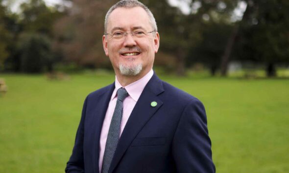 Man in a navy suit and pink shirt wearing an Irish Food Board (Bord Bia) lapel pin, standing outdoors on a green lawn with trees in the background.