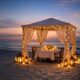 Private candlelit beachfront dining setup at Al Raha Beach Resort & Spa, featuring a draped gazebo with fairy lights, a table set for two, and lanterns on the sand at sunset overlooking the sea.