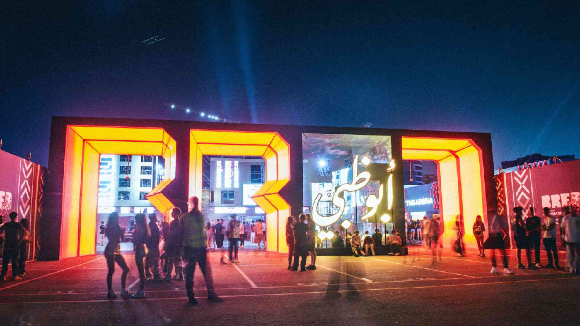 Entrance view of BRED festival at night showing illuminated orange BRED letter structures, digital Arabic calligraphy screen, and attendees walking and gathering at the venue.