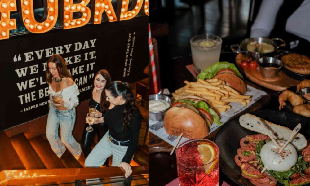 UBK venue interior showing guests socialising on a staircase beneath illuminated signage, alongside a table spread with burgers, fries, seafood dishes, and cocktails served in a lively dining atmosphere.