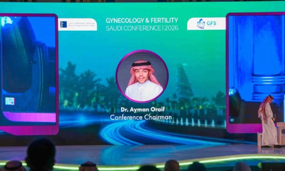 Stage view at the First Saudi Conference on Gynecology and Fertility 2026 showing a large LED screen with the event branding and Dr. Ayman Oraif identified as Conference Chairman, while an attendee in traditional Saudi attire walks across the stage and the audience is seated in front.