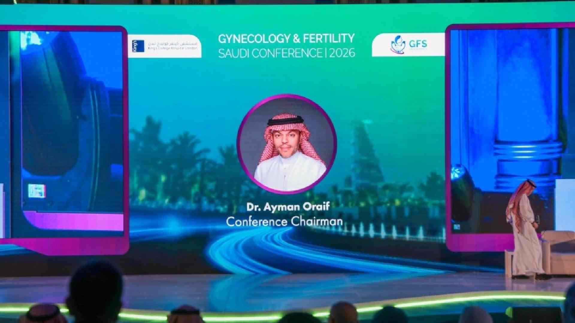 Stage view at the First Saudi Conference on Gynecology and Fertility 2026 showing a large LED screen with the event branding and Dr. Ayman Oraif identified as Conference Chairman, while an attendee in traditional Saudi attire walks across the stage and the audience is seated in front.