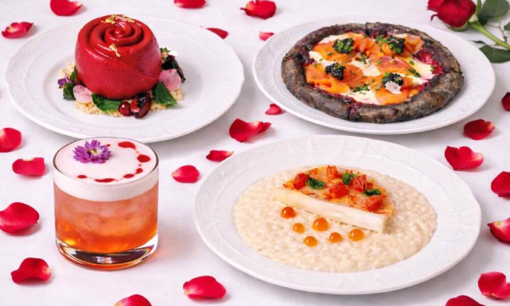 Artisan Valentine’s Day dining spread featuring dark chocolate rose dessert, salmon pizzetta with caviar, white asparagus risotto, and a cocktail, styled on a white table with scattered red rose petals