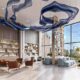 Double-height modern lobby interior by Opaal Interiors featuring floor-to-ceiling windows, sculptural blue suspended ceiling installation, built-in shelving, lounge seating, and city views beyond.