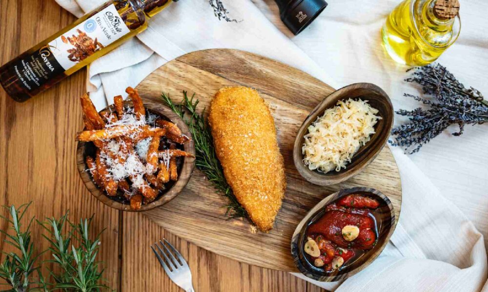 Top‑down view of a plated breaded fried cutlet with rosemary, sweet potato fries, sauerkraut, roast tomatoes, olive oil and seasonings on a wooden table at ME by Meliá Dubai