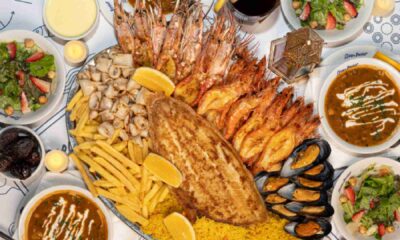 Overhead view of an Ocean Basket Ramadan seafood sharing platter with grilled fish, prawns, mussels, calamari, fries and spiced rice, surrounded by bowls of harira soup, strawberry salad, dates and drinks on a dining table.