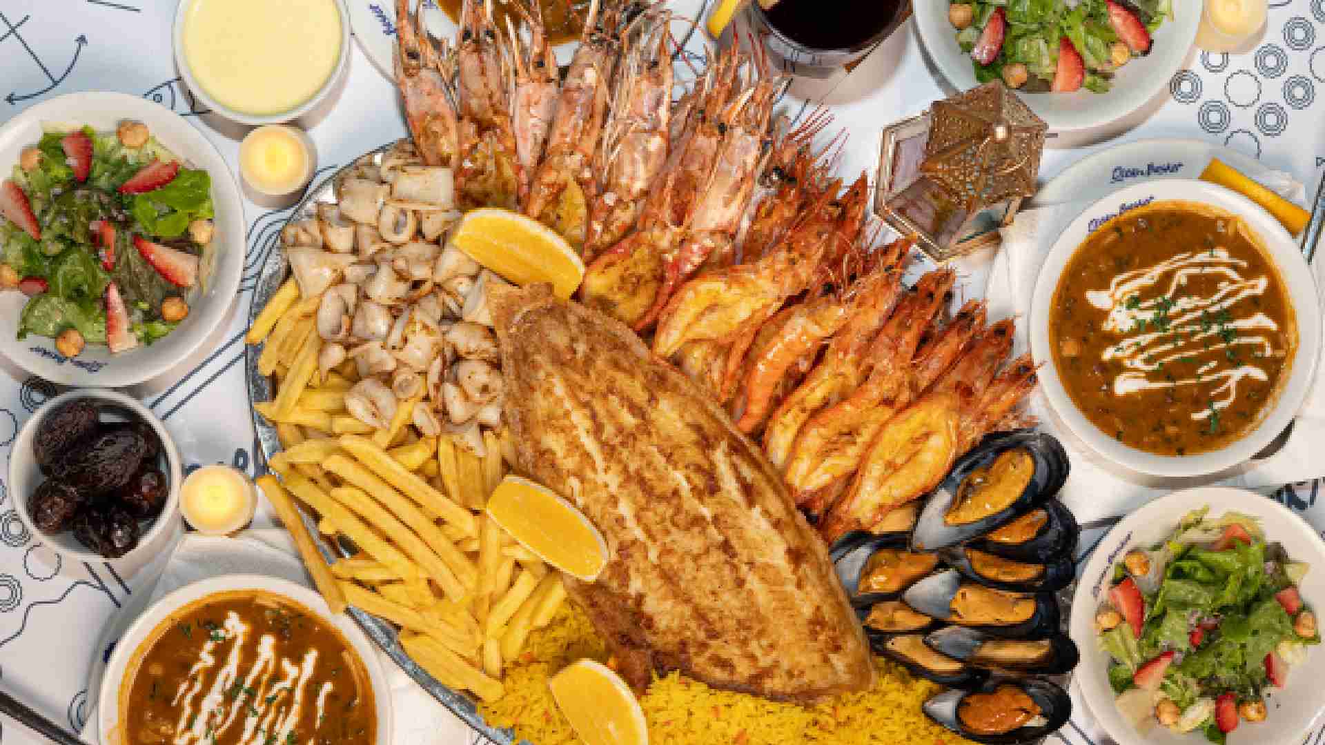 Overhead view of an Ocean Basket Ramadan seafood sharing platter with grilled fish, prawns, mussels, calamari, fries and spiced rice, surrounded by bowls of harira soup, strawberry salad, dates and drinks on a dining table.