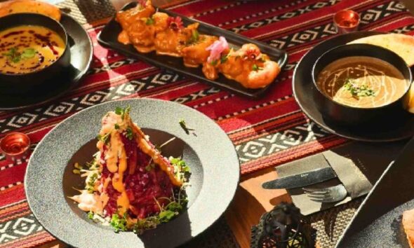 A table at JOE’S CAFÉ DUBAI set for Iftar, featuring assorted dishes including lentil soup, avocado quinoa salad, grilled skewers, a beetroot and greens starter, slow‑cooked lamb shank with rice, bread slices, and a mint drink, all arranged on a patterned red tablecloth with small lanterns and warm natural lighting.