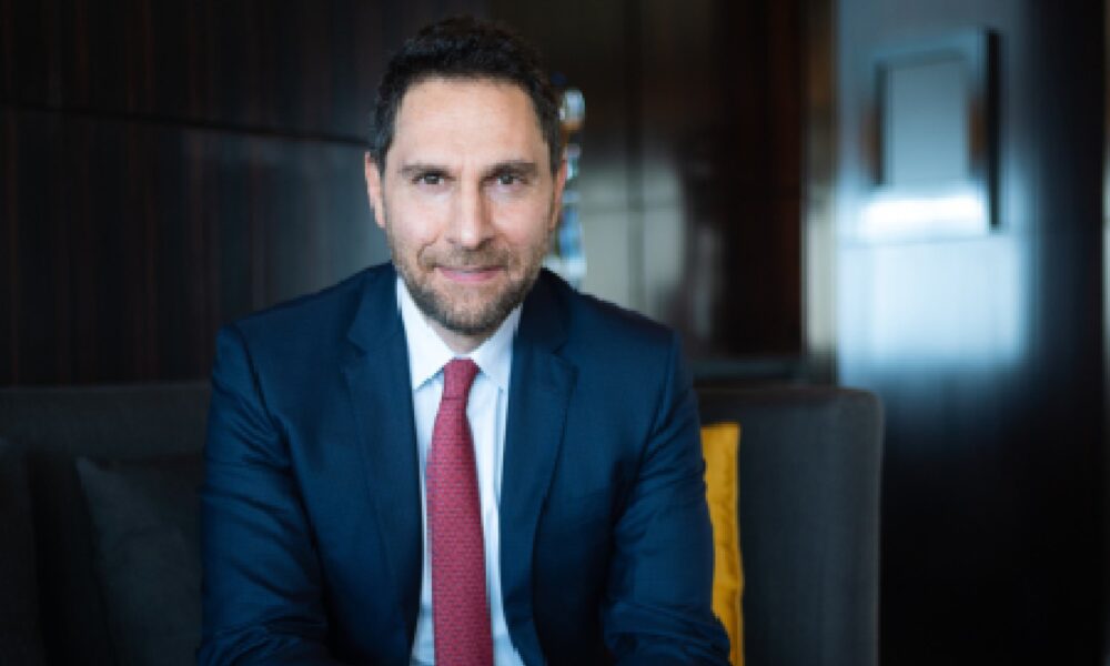 Executive from IHG Hotels & Resorts seated indoors on a sofa, wearing a navy business suit and red tie, photographed in a modern hotel lounge setting.