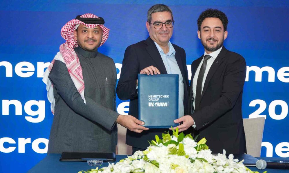 Three executives stand behind a table holding a signed document folder during a Nemetschek Group and Ingram Micro partnership announcement event, with branded backdrop and floral arrangement in front.