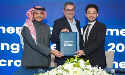 Three executives stand behind a table holding a signed document folder during a Nemetschek Group and Ingram Micro partnership announcement event, with branded backdrop and floral arrangement in front.