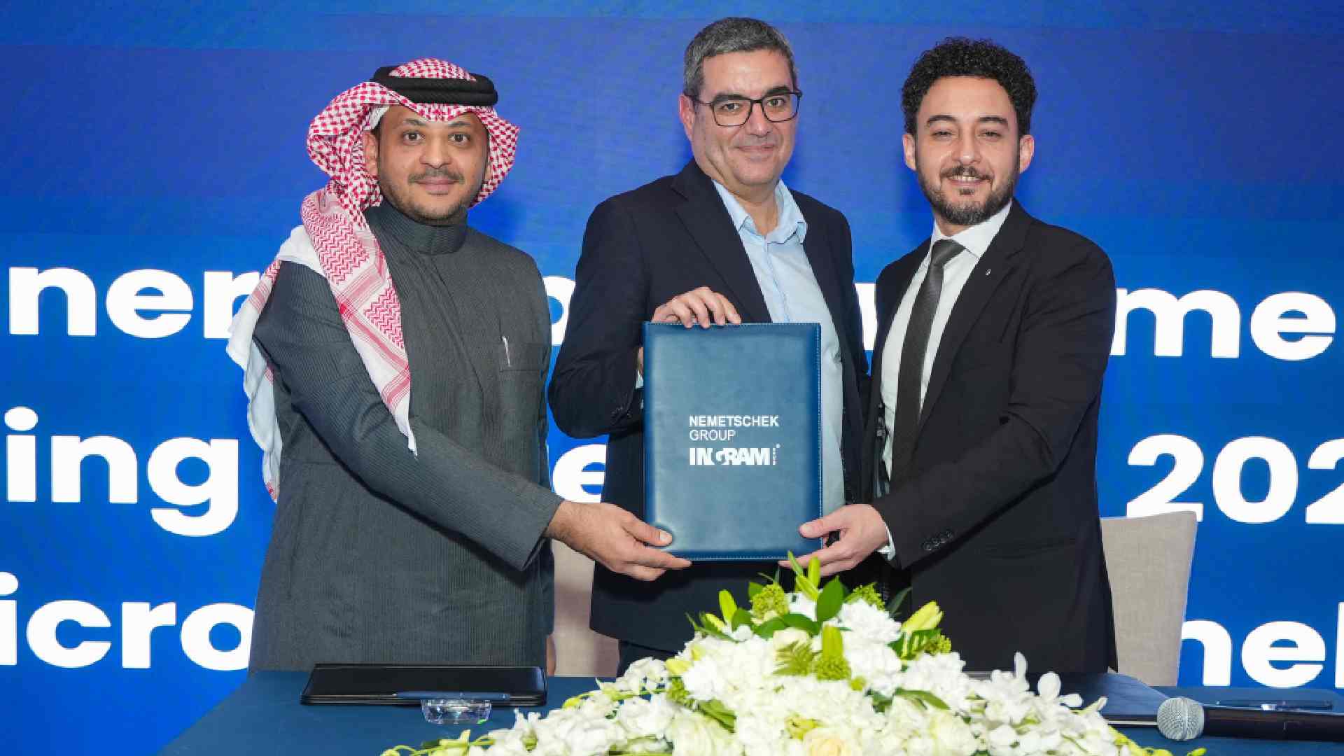 Three executives stand behind a table holding a signed document folder during a Nemetschek Group and Ingram Micro partnership announcement event, with branded backdrop and floral arrangement in front.