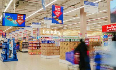 Interior view of a Carrefour supermarket showing household and grocery aisles with stacked products, hanging “Low Prices” promotional signage, and blurred shoppers pushing carts, highlighting discounted everyday essentials.