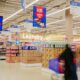 Interior view of a Carrefour supermarket showing household and grocery aisles with stacked products, hanging “Low Prices” promotional signage, and blurred shoppers pushing carts, highlighting discounted everyday essentials.