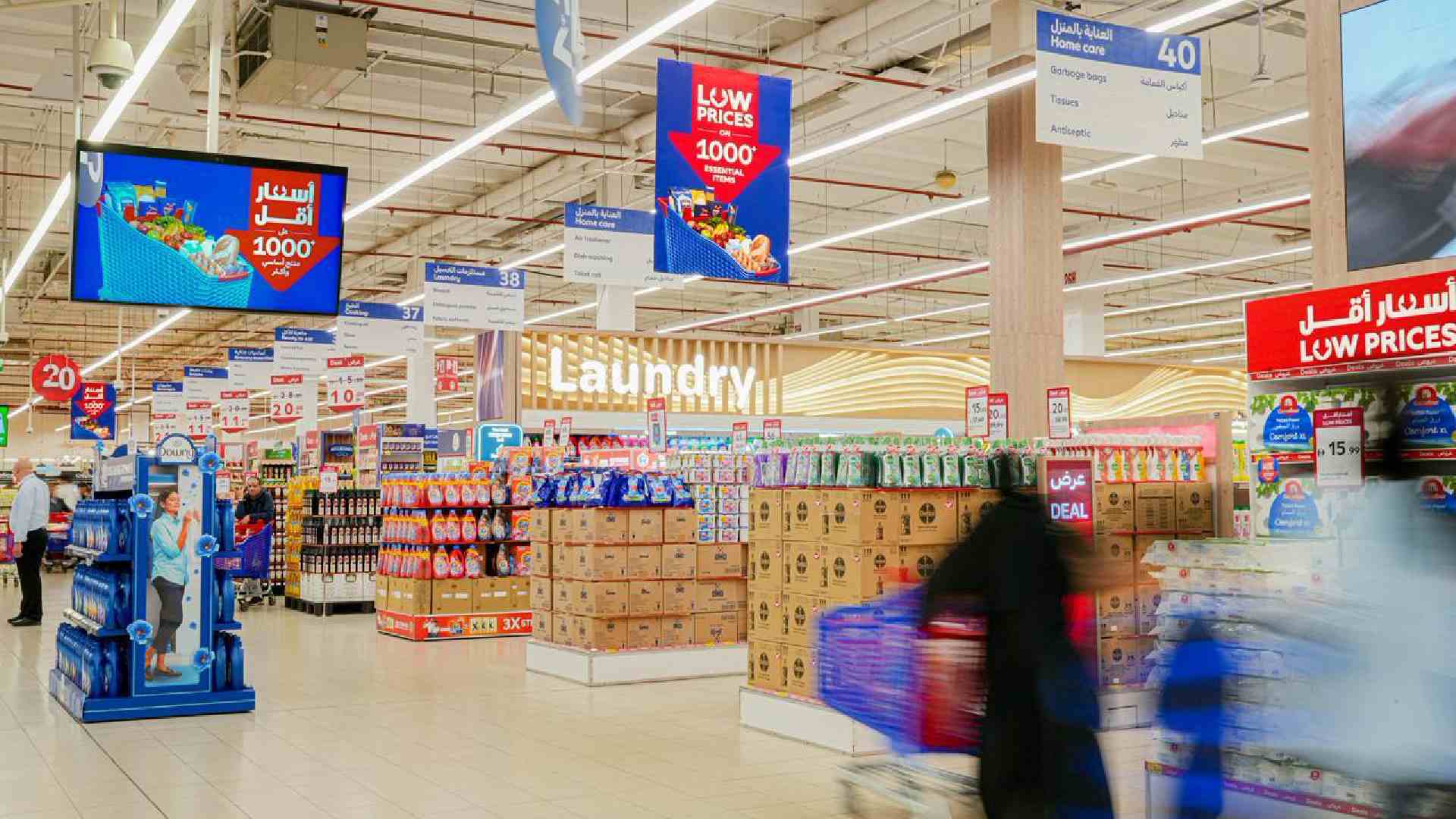 Interior view of a Carrefour supermarket showing household and grocery aisles with stacked products, hanging “Low Prices” promotional signage, and blurred shoppers pushing carts, highlighting discounted everyday essentials.