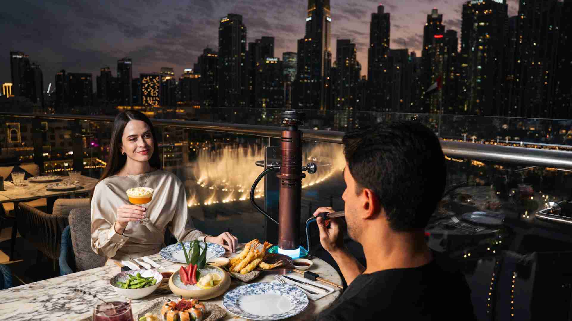 Couple dining at Blume on an outdoor terrace, sharing dessert and drinks with Dubai skyline and waterfront fountain views at night.